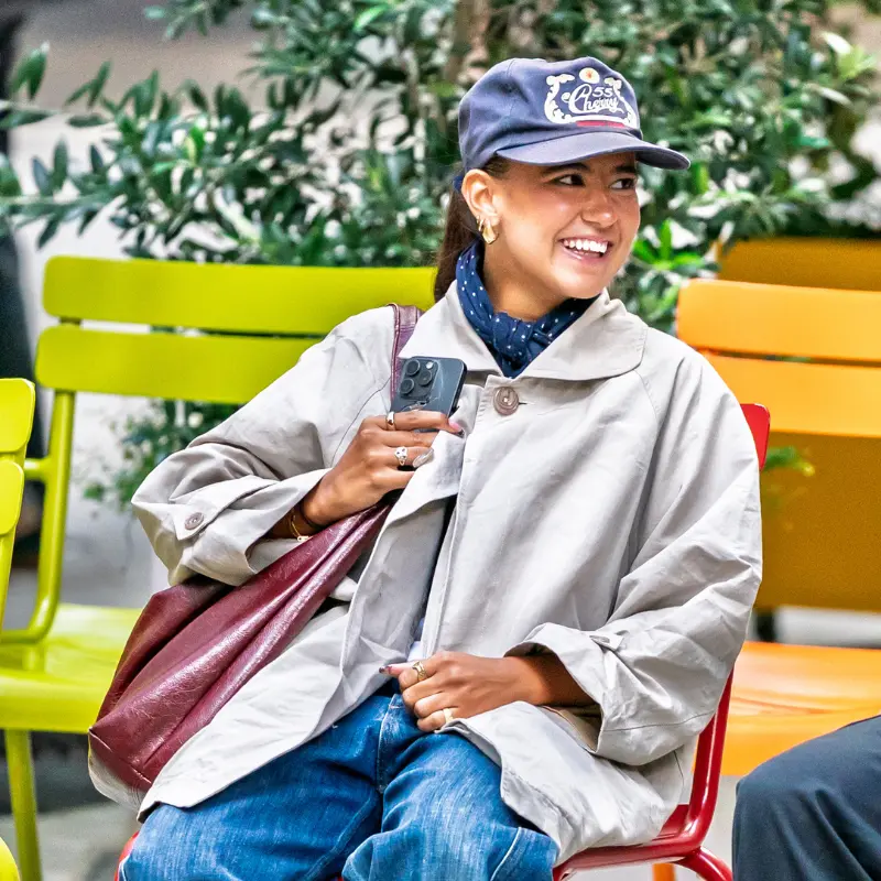 Girl wearing a cap and a burgundy bag smiling while sitting on colourful seating